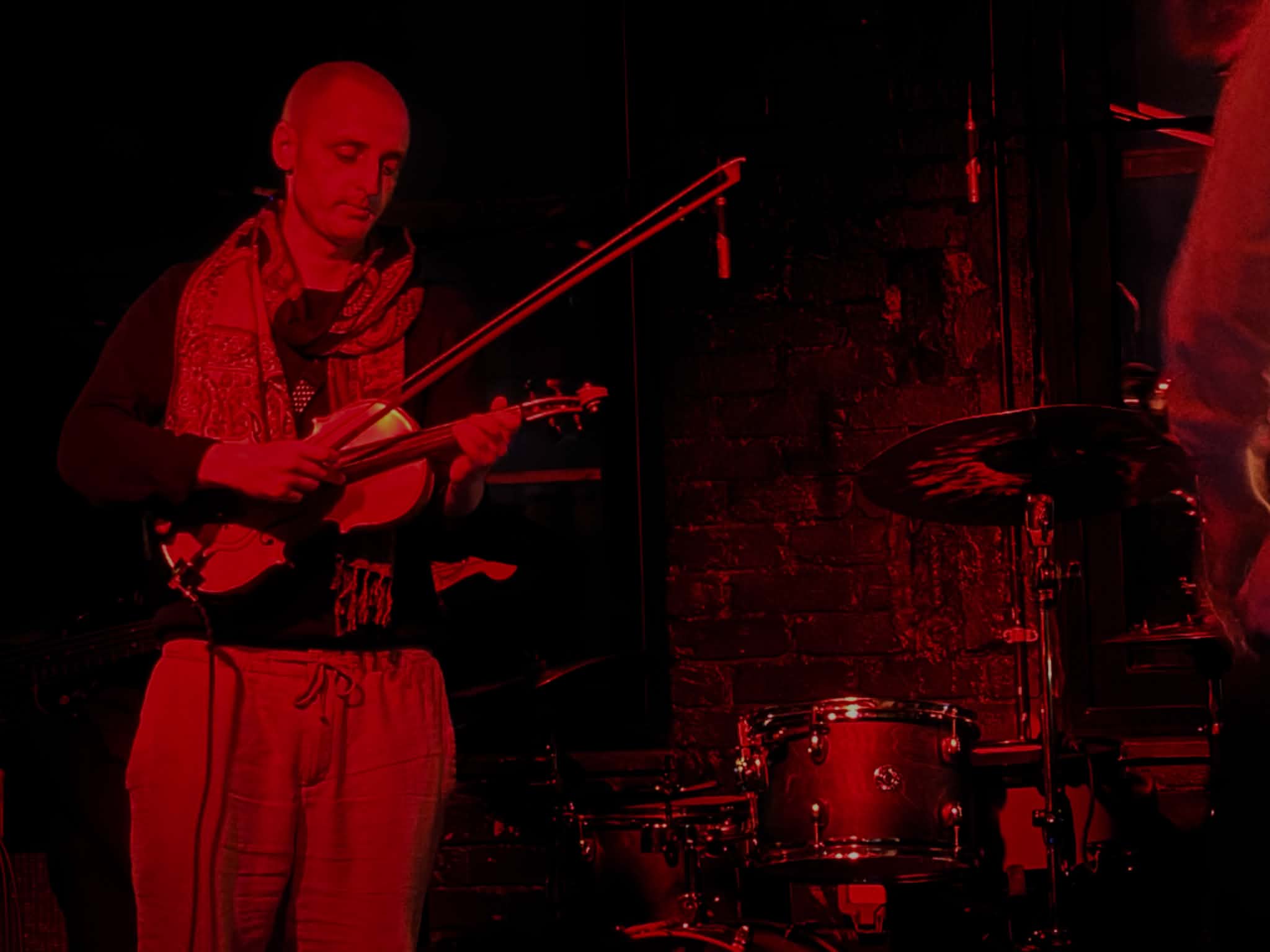 Ed White playing the violin on a dark stage lit in deep red, beside a drum kit